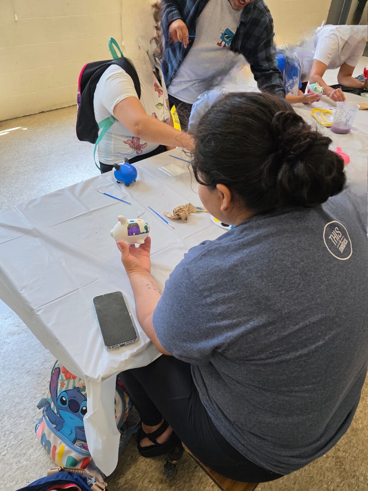 shows back of kids head and blurred pictures of 2 young adults at table painting piggy-banks