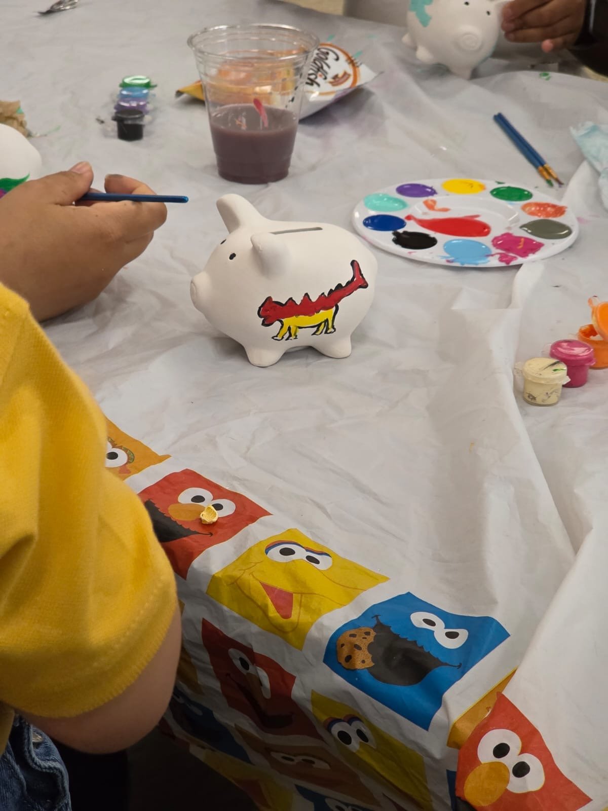 hand on table with paintbrush, piggy-bank and paint on top of white tablecloth that has pictures of sessame street characters