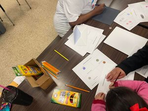 2 kids sitting diagonally across from one another, with paper, pencils and crayons on the table