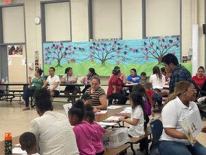 woman holding open a book telling a story, with back to classroom with adults and kids
