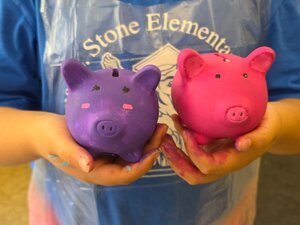 student showing 2 painted piggy banks, one in each hand, shows student from chest to end of torso